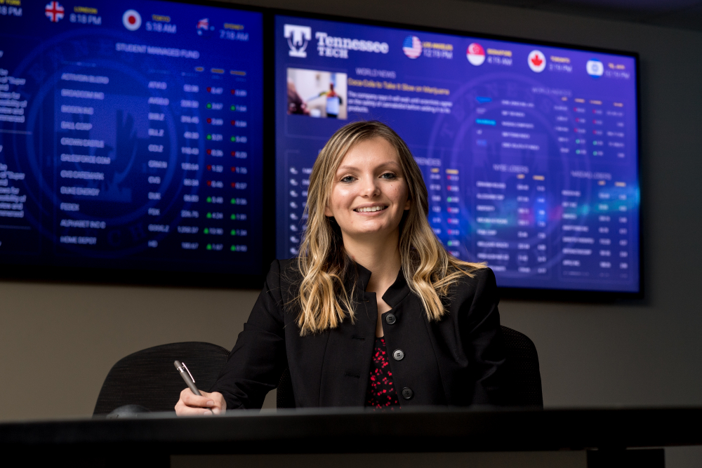 smiling Student behind desk with TV in background. 